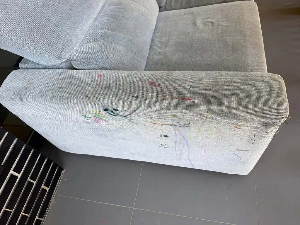 Detalle del lateral de un sofá de tela gris claro lleno de trazos de rotulador antes de la limpieza